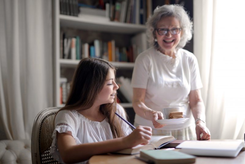 Older woman giving cookies and tea to younger person, doing homework at home